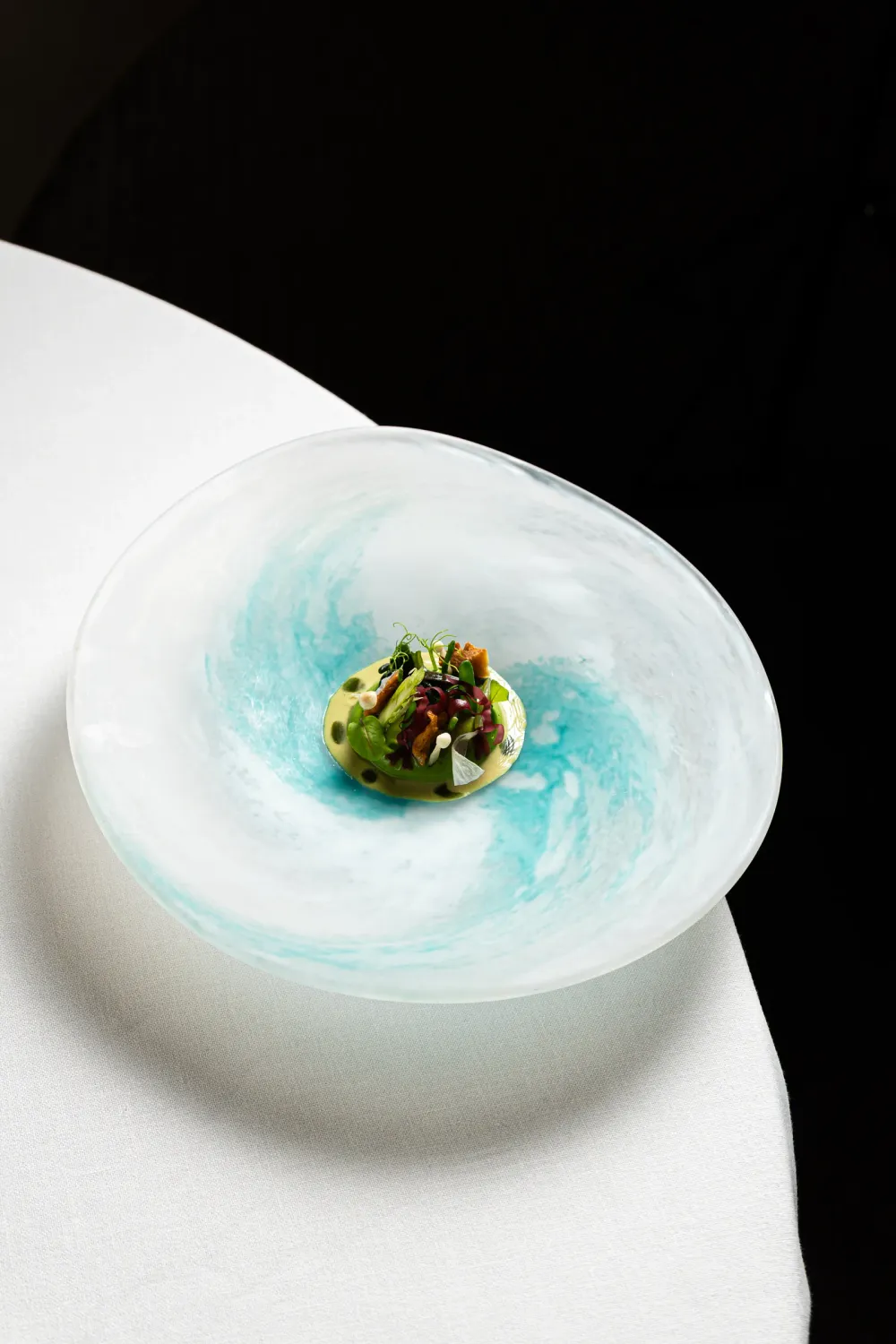 Plato de cristal azul turquesa con una ensalada de verduras y setas sobre una mesa con mantel blanco y fondo negro